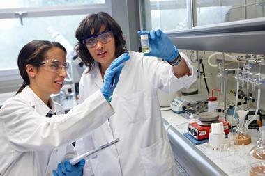 Two women wearing white lab coats, goggles and blue nitrile gloves look at a vial being held up by the person on the right. They are stood in a lab next to a fume hood